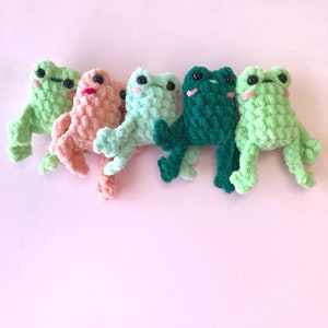 Op de afbeelding: Vijf gehaakte kikker knuffels in verschillende kleuren: groen, lichtgroen, perzik en donkergroen. De kikkers kijken allemaal naar voren met hun ogen dicht.