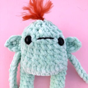 May include: A crocheted plush toy with a green body, black eyes, and a red yarn mohawk. The toy has a simple, friendly expression.