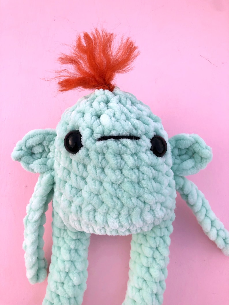 May include: A crocheted plush toy with a green body, black eyes, and a red yarn mohawk. The toy has a simple, friendly expression.