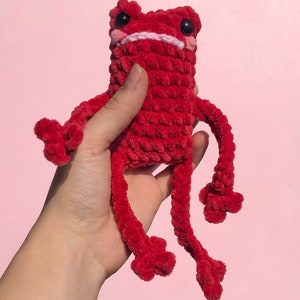 Op de afbeelding: Een rood gehaakt knuffeldier in de vorm van een kikker met zwarte ogen en een roze mond. De kikker heeft lange, dunne armen en benen.
