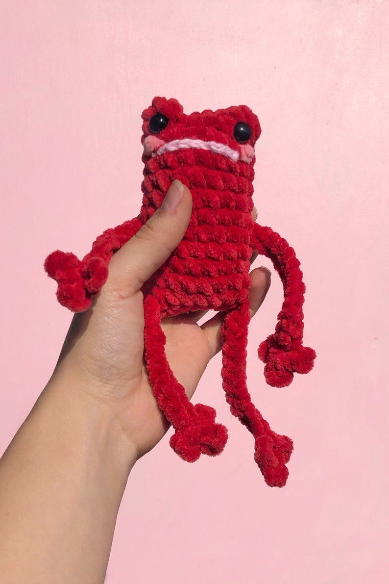 Op de afbeelding: Een rood gehaakt knuffeldier in de vorm van een kikker met zwarte ogen en een roze mond. De kikker heeft lange, dunne armen en benen.