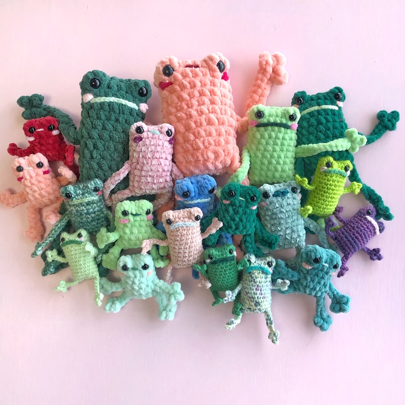 Op de afbeelding: Een verzameling gehaakte kikker knuffels in verschillende kleuren, waaronder groen, blauw, roze en oranje. De kikkers zijn allemaal van verschillende grootte en hebben verschillende gezichtsuitdrukkingen.