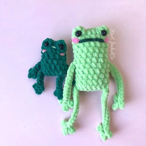 Op de afbeelding: Twee gehaakte kikker knuffels, een groene en een donkergroene, met zwarte ogen en roze wangen. De grotere kikker heeft een zwarte mond.