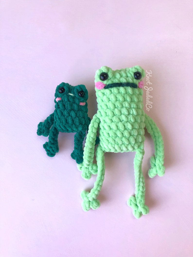 Op de afbeelding: Twee gehaakte kikker knuffels, een groene en een donkergroene, met zwarte ogen en roze wangen. De grotere kikker heeft een zwarte mond.
