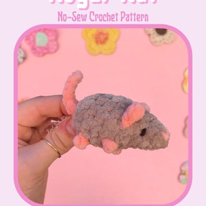 May include: A gray and pink crocheted rat named Roger Rat. The crochet pattern is available for purchase. The image is on a pink background with crocheted flowers.