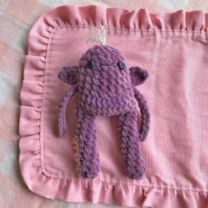 May include: Purple crocheted plush toy with long arms and legs, sitting on a pink ruffled blanket. The toy has a friendly face with black eyes and a small mouth. The blanket has the text "Knot Jaded Co." embroidered on it.