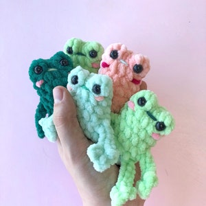 Op de afbeelding: Vijf gehaakte kikker knuffels in verschillende kleuren, waaronder groen, lichtgroen en roze. De knuffels hebben grote ogen en worden in een hand gehouden.