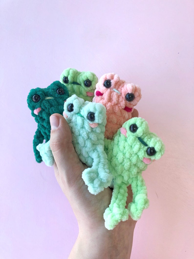Op de afbeelding: Vijf gehaakte kikker knuffels in verschillende kleuren, waaronder groen, lichtgroen en roze. De knuffels hebben grote ogen en worden in een hand gehouden.