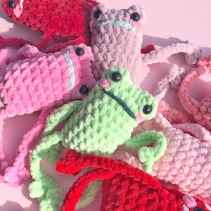 Op de afbeelding: Een groep van vijf gehaakte kikker knuffels in verschillende kleuren, waaronder rood, roze, groen en paars. De kikkers hebben grote, zwarte ogen en zijn gemaakt van pluche garen.