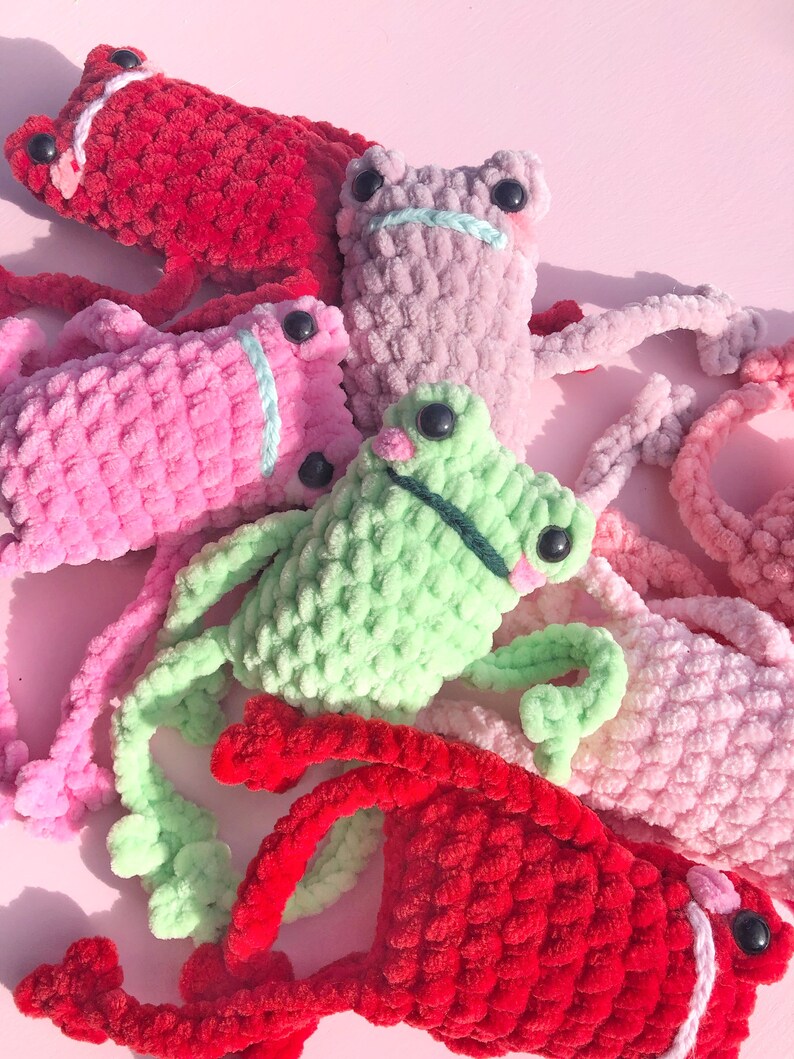 Op de afbeelding: Een groep van vijf gehaakte kikker knuffels in verschillende kleuren, waaronder rood, roze, groen en paars. De kikkers hebben grote, zwarte ogen en zijn gemaakt van pluche garen.
