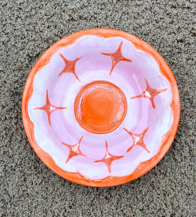 May include: A small, orange ceramic dish with a white center and a pink rim. The dish has a scalloped edge and is decorated with six star-shaped designs.