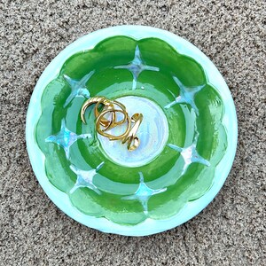 May include: A light blue and green ceramic trinket dish with a starburst design. The dish is filled with gold rings.
