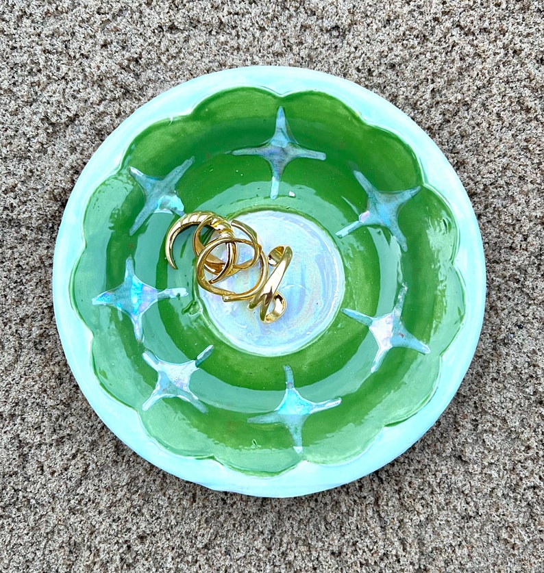May include: A light blue and green ceramic trinket dish with a starburst design. The dish is filled with gold rings.