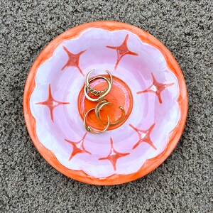 May include: A pink and orange ceramic dish with a scalloped edge and a starburst design. The dish is filled with gold rings.