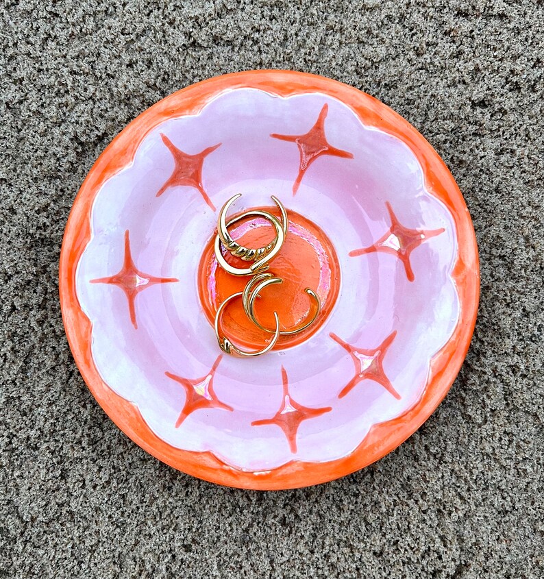May include: A pink and orange ceramic dish with a scalloped edge and a starburst design. The dish is filled with gold rings.