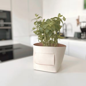 Op de afbeelding: Een crèmekleurige leren plantenbak met een rechthoekige patch, met een groene muntplant. De binnenkant van de plantenbak is lichtbruin. Hij staat op een wit oppervlak, waarschijnlijk in een keuken.