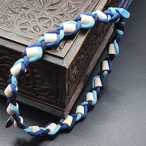 May include: A blue and white woven necklace with rectangular white beads. The necklace is made of blue and light blue cord, with a clasp and a blue button closure. The necklace is displayed on a dark wooden surface.