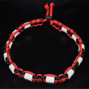 May include: A woven paracord bracelet in red, black, and white. The bracelet has a distinctive design with alternating colours and white cylindrical beads. The adjustable closure is red.