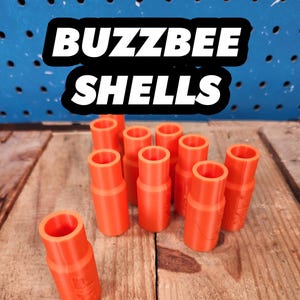 May include: Orange "Buzzbee Shells" are displayed on a wooden surface. The cylindrical shells are arranged in a group, with the text "BUZZBEE SHELLS" in a black and white banner above them. The shells are made of plastic.