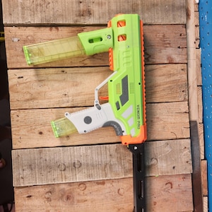 K&ouml;nnte beinhalten: Eine gr&uuml;ne und orangefarbene Spielzeugpistole mit dem Text "VENOM PRO" wird auf einer Holzoberfl&auml;che gezeigt. Die Spielzeugpistole hat ein transparentes Magazin und einen schwarzen, verstellbaren Schaft. Die Pistole ist etwa 71 cm lang.