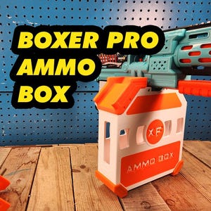 May include: A white and orange ammo box with the text "BOXER PRO AMMO BOX" in yellow and black. The ammo box is attached to a teal and orange toy gun. The background is a blue pegboard.