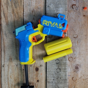 May include: A blue and yellow toy gun with the word "Rival" printed on it. The gun is disassembled and the barrel is separate from the main body.