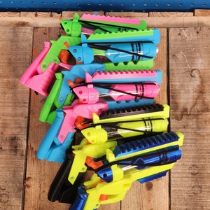May include: A set of seven colorful toy guns with black elastic bands and clear plastic tubes. The guns are made of plastic and have a variety of colors, including pink, blue, green, and yellow.
