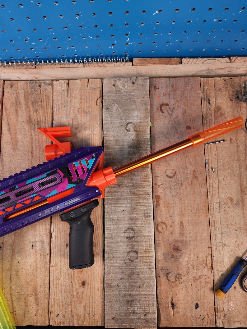 Puede incluir: Una pistola de juguete morada y naranja con una empu&ntilde;adura negra y un ca&ntilde;&oacute;n naranja. La pistola est&aacute; sobre una superficie de madera.