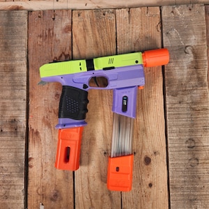 May include: A purple and green toy gun with an orange barrel and a clear magazine. The gun has a black grip and a textured surface.