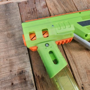 K&ouml;nnte beinhalten: Eine gr&uuml;ne und orangefarbene Spielzeugpistole mit einem transparenten Magazin. Die Spielzeugpistole hat ein rechteckiges Design mit Griff und Abzug. Die Pistole ist aus Kunststoff und hat ein paar sichtbare Schrauben.