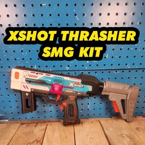 May include: A toy blaster labeled XSHOT THRASHER SMG KIT featuring a white, teal, and black color scheme with orange accents. The blaster includes a textured gray stock, a black grip, and a red trigger, with various geometric markings and text.