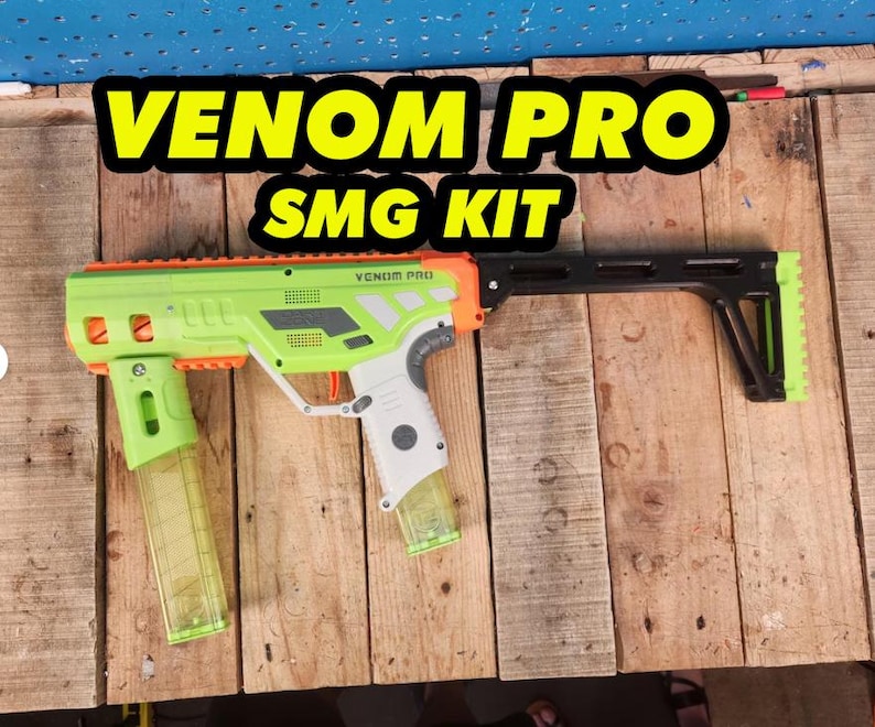 K&ouml;nnte beinhalten: Eine gr&uuml;ne und orangefarbene Spielzeugpistole mit dem Text "VENOM PRO SMG KIT" in Gelb und Schwarz. Die Spielzeugpistole hat einen schwarzen Schaft und ein transparentes Magazin.
