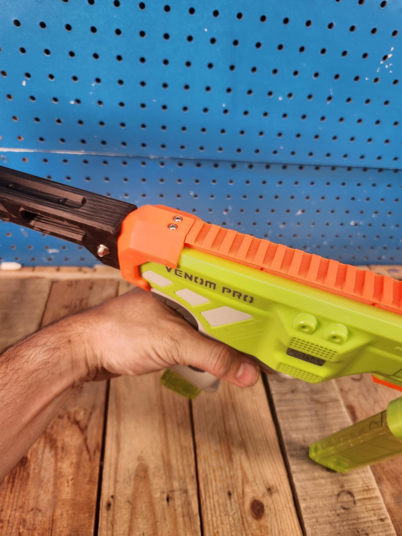 K&ouml;nnte beinhalten: Eine neongr&uuml;ne und orangefarbene Spielzeugpistole mit dem Text "VENOM PRO" auf der Seite. Die Spielzeugpistole hat einen schwarzen Lauf und wird von einer Person gehalten.