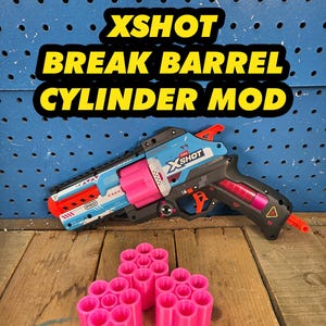 May include: A Zuru X-Shot toy blaster with a blue, white, and black body, an orange trigger, and a bright pink rotating cylinder. Three additional bright pink cylindrical components are positioned in front of the blaster. Text reads XSHOT BREAK BARREL CYLINDER MOD.