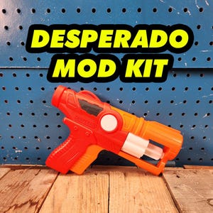 May include: A red and orange toy gun with the words "Adventure Strike" and "Desperado" printed on it. The gun is on a wooden surface.