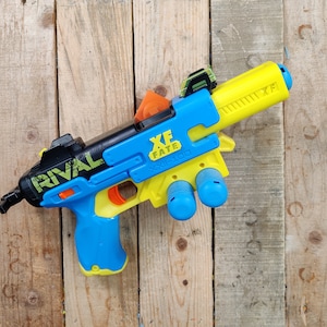 May include: A blue and yellow toy blaster with a black barrel and a green and yellow "RIVAL" logo. The blaster has a blue handle and a yellow trigger. The blaster has a black and yellow "XF FATE XXI-100" label on the side.