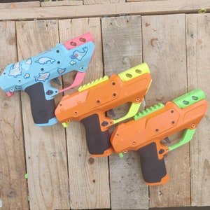 May include: Three toy water guns in different colors. The first gun is light blue with a white and light blue design. The second gun is light green and yellow. The third gun is orange with a green top.