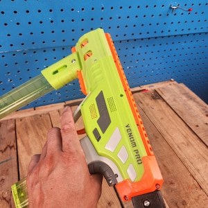 K&ouml;nnte beinhalten: Eine neongr&uuml;ne und orangefarbene Spielzeugpistole mit dem Text "VENOM PRO" an der Seite. Die Spielzeugpistole hat ein transparentes Magazin und wird von einer Hand gehalten. Der Hintergrund ist eine blaue Lochwand und Holzbretter.