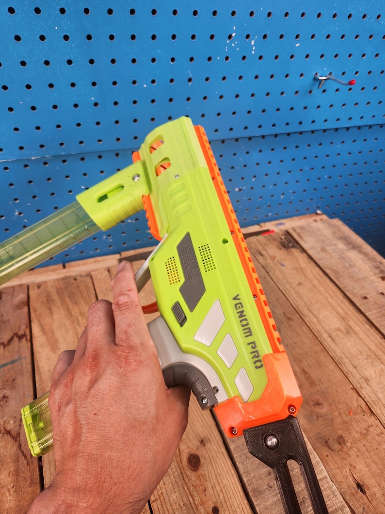 K&ouml;nnte beinhalten: Eine neongr&uuml;ne und orangefarbene Spielzeugpistole mit dem Text "VENOM PRO" an der Seite. Die Spielzeugpistole hat ein transparentes Magazin und wird von einer Hand gehalten. Der Hintergrund ist eine blaue Lochwand und Holzbretter.