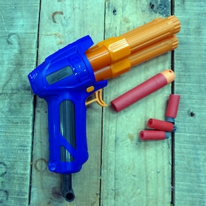 May include: A blue and orange toy gun with a black grip and a barrel that holds multiple orange darts. Four red darts are lying on a wooden surface.
