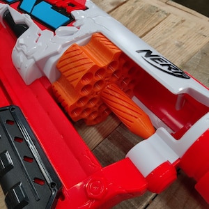 May include: A red and white Nerf toy gun with an orange clip. The clip is visible and has a large number of darts.