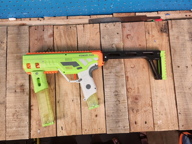 K&ouml;nnte beinhalten: Eine hellgr&uuml;ne und orangefarbene Spielzeugpistole mit dem Text "VENOM PRO" auf der Seite. Die Spielzeugpistole hat einen schwarzen Schaft und zwei transparente Magazine. Die Spielzeugpistole liegt auf einer Holzoberfl&auml;che.