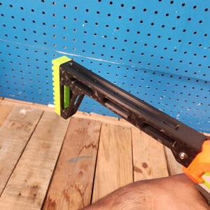 K&ouml;nnte beinhalten: Schwarzer und neongr&uuml;ner Nerf-Gewehrschaftaufsatz. Der Aufsatz besteht aus Kunststoff und hat ein strukturiertes Design. Der Schaft wird von einer Hand gehalten. Der Hintergrund ist eine blaue Lochwand und Holzbretter.