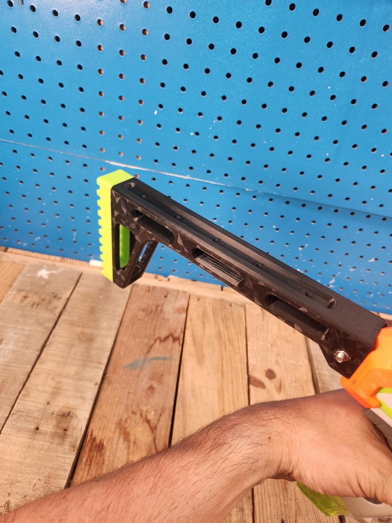 K&ouml;nnte beinhalten: Schwarzer und neongr&uuml;ner Nerf-Gewehrschaftaufsatz. Der Aufsatz besteht aus Kunststoff und hat ein strukturiertes Design. Der Schaft wird von einer Hand gehalten. Der Hintergrund ist eine blaue Lochwand und Holzbretter.