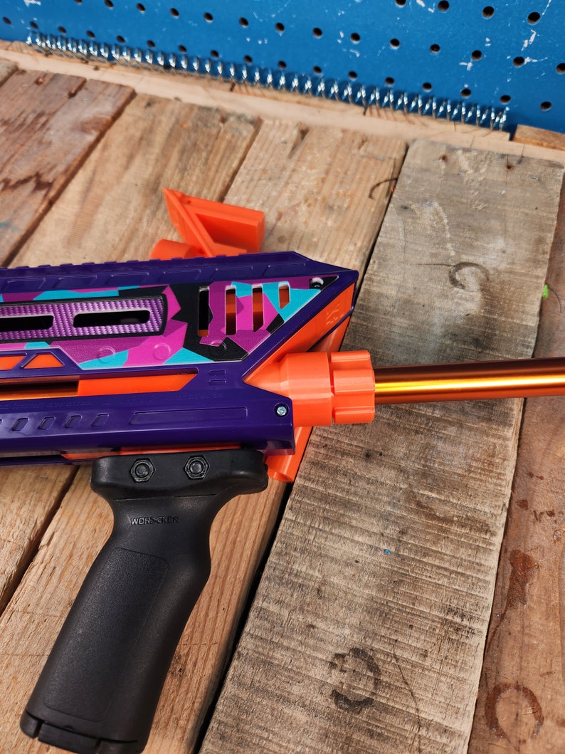 Puede incluir: Una pistola de juguete morada y naranja con una empu&ntilde;adura negra y un patr&oacute;n de camuflaje. La pistola tiene una extensi&oacute;n de ca&ntilde;&oacute;n y una empu&ntilde;adura t&aacute;ctica.