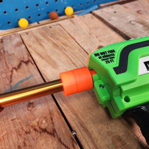 May include: A green and black toy gun with an orange barrel and a gold-colored tube. The text "DO NOT FIRE at objects closer than 25 FT" is printed on the side. Three yellow and one brown ball are on a wooden surface.