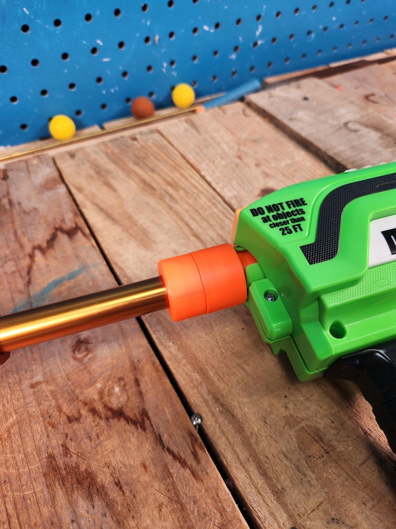 May include: A green and black toy gun with an orange barrel and a gold-colored tube. The text "DO NOT FIRE at objects closer than 25 FT" is printed on the side. Three yellow and one brown ball are on a wooden surface.