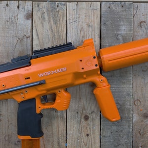 May include: An orange and black toy gun with a black barrel and a black handle. The gun has a black scope and the words "WORXCKER" printed on the side.