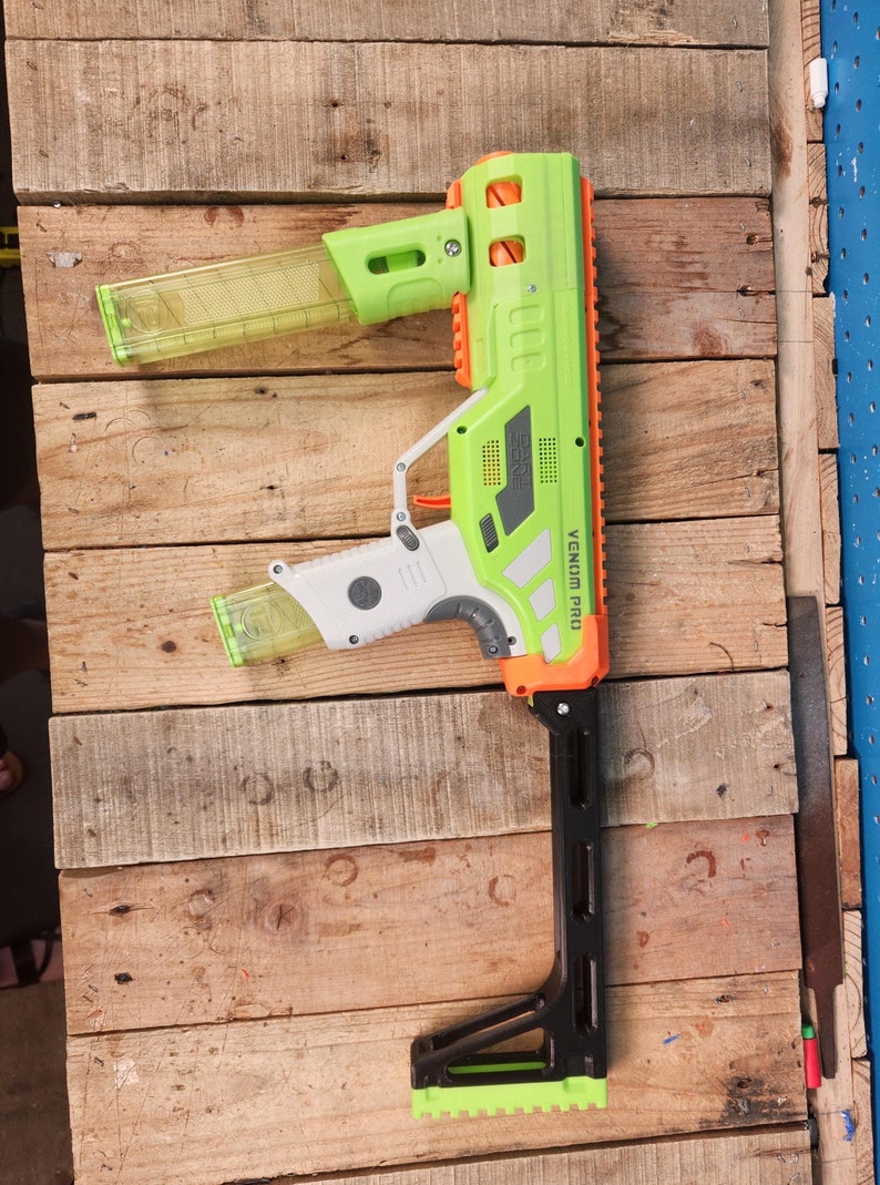 K&ouml;nnte beinhalten: Eine gr&uuml;ne und orangefarbene Spielzeugpistole mit dem Text "VENOM PRO" wird auf einer Holzoberfl&auml;che gezeigt. Die Spielzeugpistole hat ein transparentes Magazin und einen schwarzen, verstellbaren Schaft. Die Pistole ist etwa 71 cm lang.