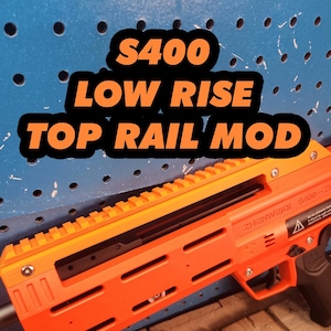 May include: Bright orange S400 Low Rise Top Rail Mod. The text "S400 LOW RISE TOP RAIL MOD" is in the image. The rail mod has a series of horizontal slots and a ridged top. The background is a blue pegboard.